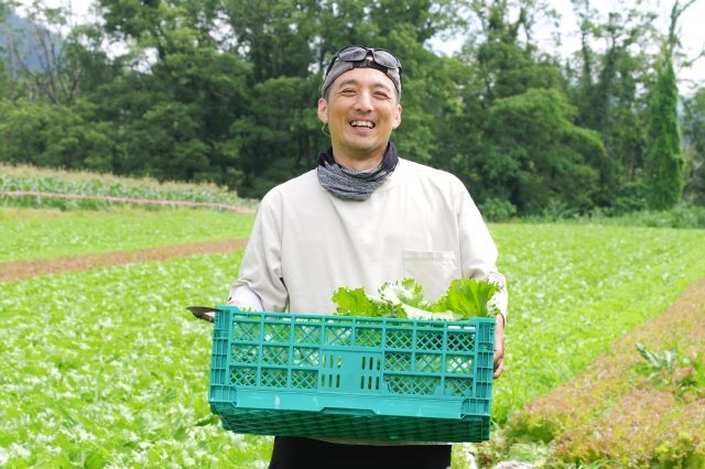 畑で収穫した野菜のケースを抱えて笑う生産者